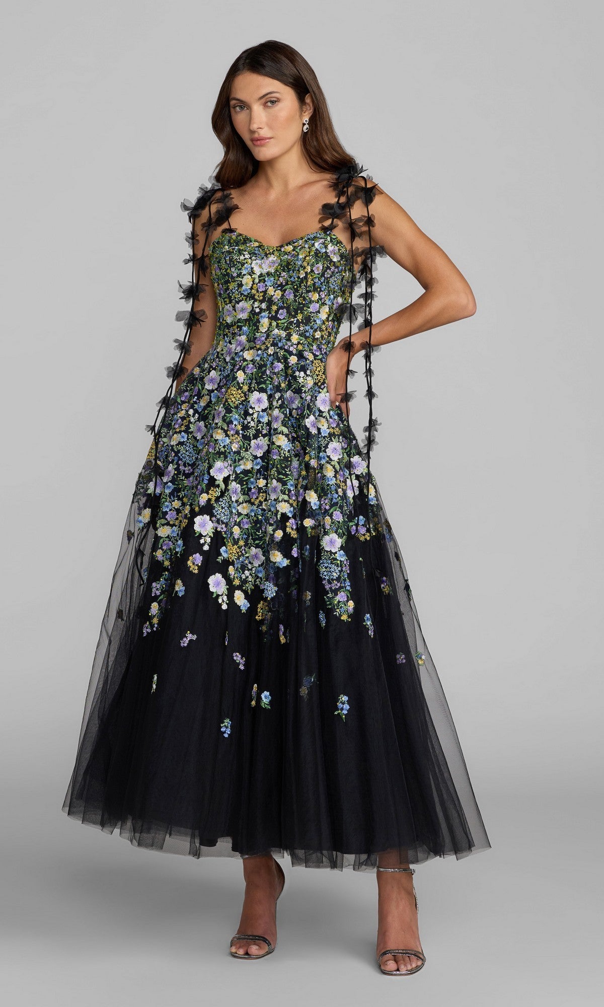 A woman wearing the Midi Party Dress: Mac Duggal 21356 in Black Multi, a black tulle midi dress adorned with multicolored floral embroidery, featuring thin straps and an elegant A-line silhouette, paired with open-toe heels and standing against a plain gray background.